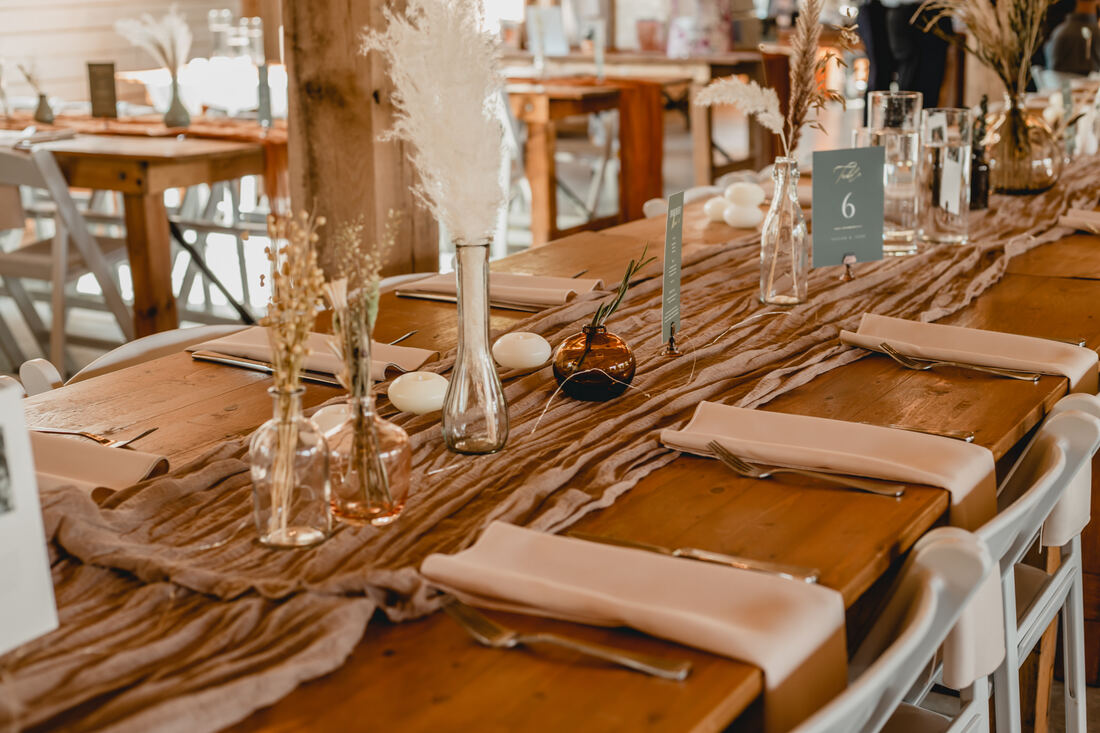 Feast wooden tables with cheesecloth runner and various vases of dried florals and pampas